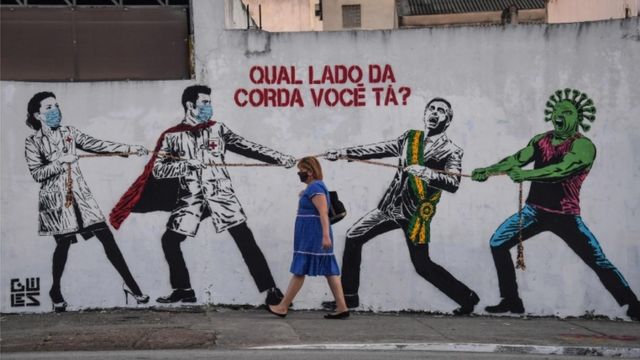 A woman passes by a graffiti depicting Brazilian President Jair Bolsonaro and a figure representing the novel coronavirus COVID-19 pulling a rope against health workers with the question "Which side of the rope are you on?" in Sao Paulo, Brazil