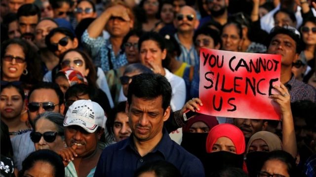 Dezenas de pessoas em protesto; duas mulheres erguem cartaz que diz em inglês: 'Você não pode nos silenciar'