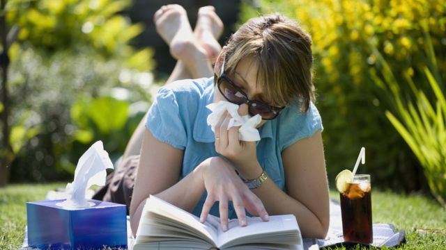 Mulher de óculos escuro assoando o nariz enquanto lê livro deitada no parque durante o verão