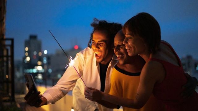Trs pessoas de cor negra celebram chegada do Ano Novo; uma delas segura vela vulco e a outra faz selfie com celular; todas riem para foto