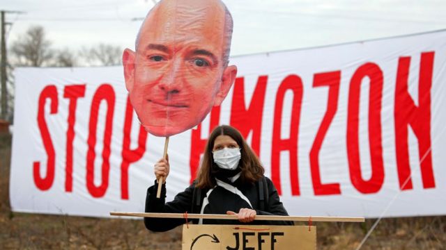 إحدى المعارضات تحمل لافتة تصور رئيس مجلس إدارة أمازون جيف بيزوس، أمام لافتة كتب عليها "أوقفوا أمازون" أثناء مسيرة ضد خطط بناء مستودع عملاق للشركة في جنوب فرنسا