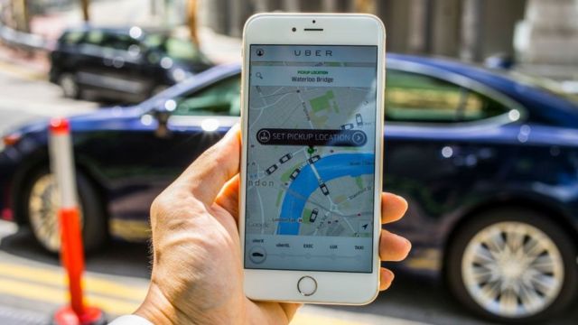 Mão segura celular com o aplicativo Uber na tela, visto de dentro de um carro