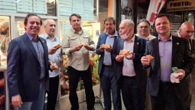 Sete homens comendo pizza em pé em frente a restaurante