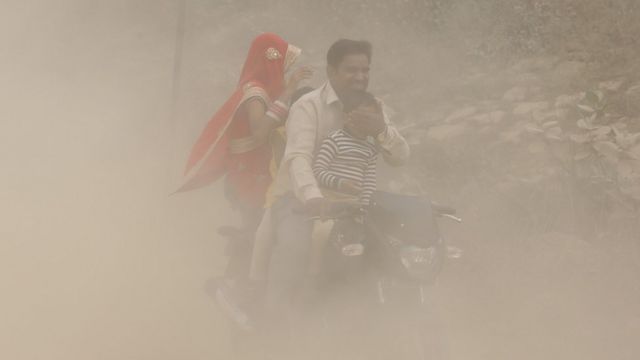 Pai, mãe e filhos em moto em meio a nuvem de poeira