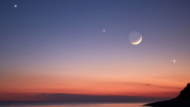 A crescent moon and three other illuminated bodies form a line over a body of water and a cliff
