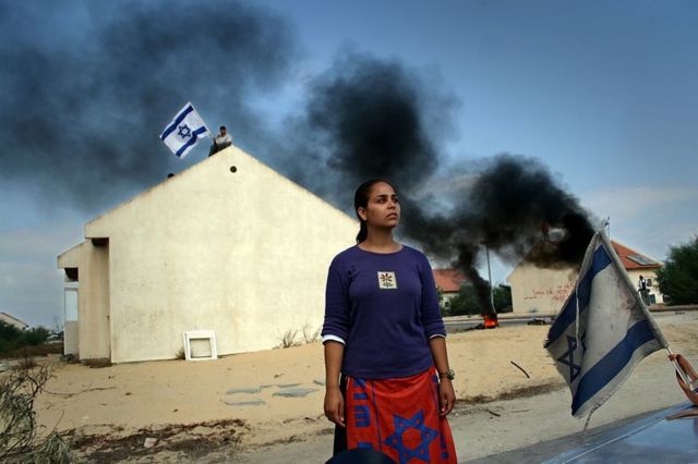 مستوطنة يهودية تلقى نظرة أخيرة على مستوطنة غوش قطيف قبل أن تغادرها عام 2005. وفي الخلفية مستوطن آخر يرفع علم إسرائيل فوق أحد مباني المستوطنة.