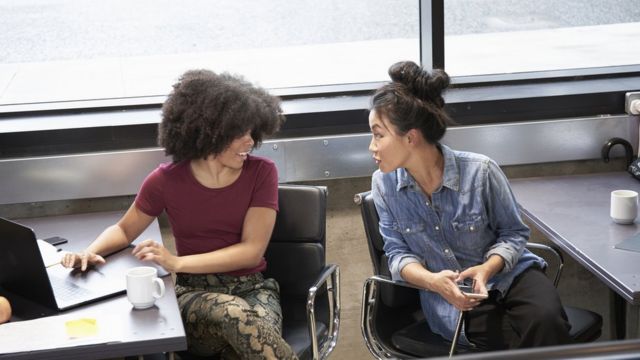 Dos mujeres conversando una con la otra, cada una su escritorio.