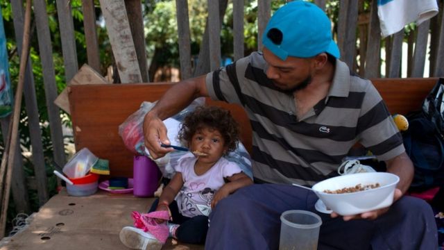 Migrante venezolano alimenta a su hija en la calle