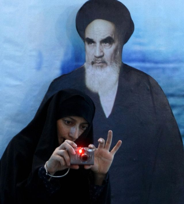 A woman poses in front of a photo of Khomeini