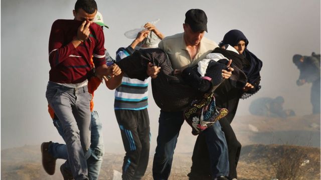 People run through tear gas carrying an injured woman at the border fence between Gaza City and Israel