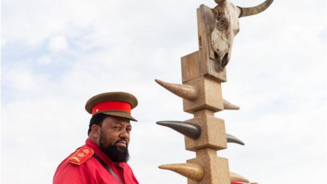 Ein hochrangiger Häuptling in traditioneller Heldenkleidung steht an einem Denkmal zu Ehren von OvaHerero und den Bewohnern des Hauses, die Anfang des 20.