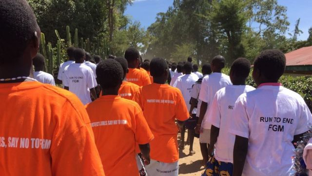 Students walk in a line wearing orange t-shirts that say "Protect girls, say no to FGM" and white t-shirts saying: Run To end FGM