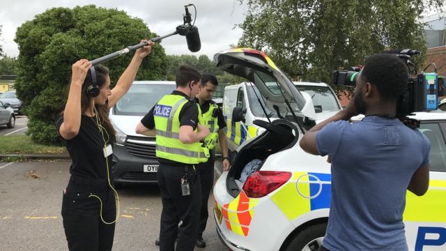 A camera operator and a sound recordist with a boom mic record two police officers standing by a police car