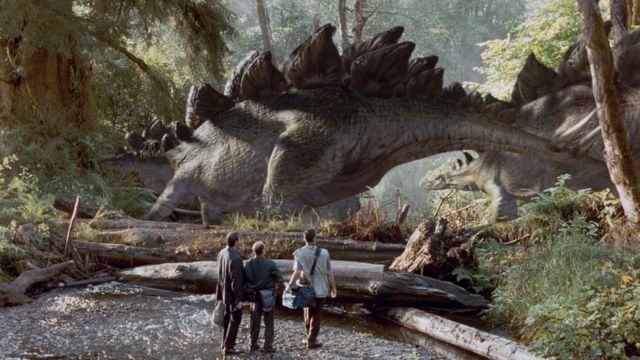Cena de filme mostra um dinossauro enorme com espinhos nas costas sendo observado por humanos