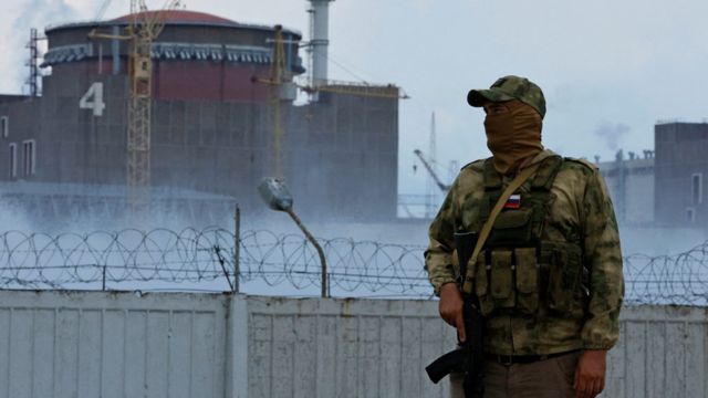 Fotografia colorida mostra um soldado russo com roupas de estampa camuflada e rosto coberto do lado de fora de uma cerca que guarda a usina de Zaporizhzhia em agosto de 2022/ uma grande estrutura redonda vermelha pode ser vista ao fundo
