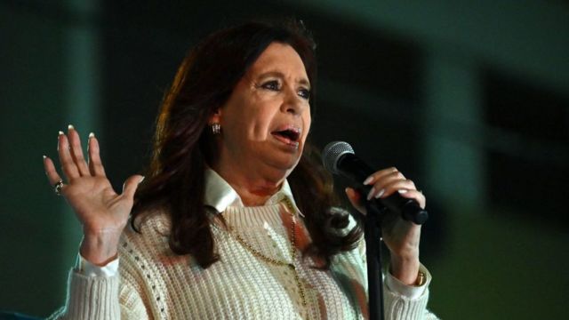 Cristina, con un micrófono en la mano, la otra palma abierta, mientras habla, vestida con una camisa blanca y un suéter