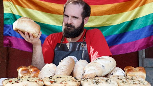 Gabriel Castro segura um pão com a mão direita, sentado em uma mesa cheia de diferentes tipos de pães, com uma bandeira de arco-íris ao fundo