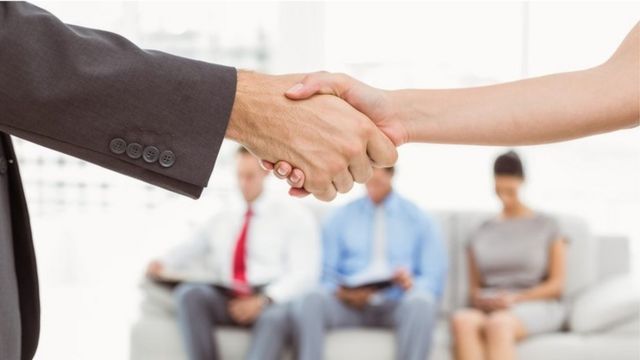 Dos personas dándose la mano. Al fondo, tres personas esperan turno para una entrevista de trabajo