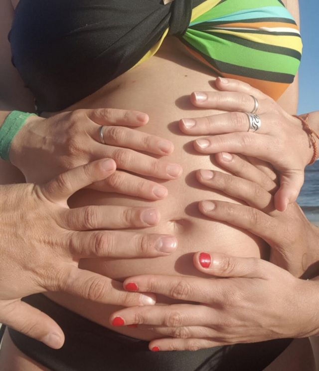 Imagem mostra várias mãos sobre a barriga de uma mulher grávida