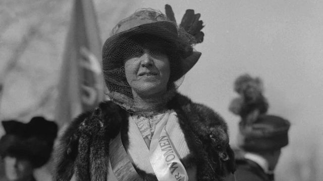 Katharine McCormick portant une écharpe sur laquelle figure le mot "Women", avec une bannière tenue en arrière-plan.