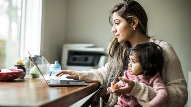 Stock image of a mum holding an infant while working on a laptop.