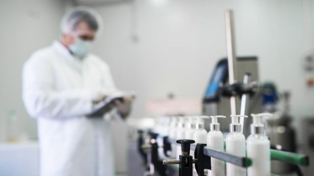 Frascos de creme em close em um laboratório; ao fundo homem com jaleco branco, máscara e protetor de cabelo olha algo em uma tele