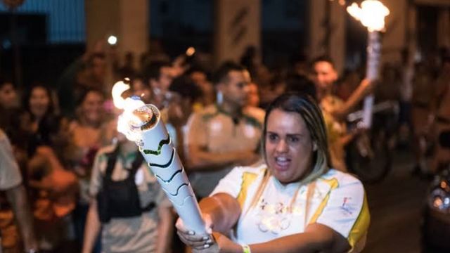 Bianka Acsa Rosa da Fonseca segura com as duas mãos a tocha olímpica flamejante em frente ao corpo, vestida com o uniforme branco e amarelo usado pelos condutores nas Olimpíadas de 2016