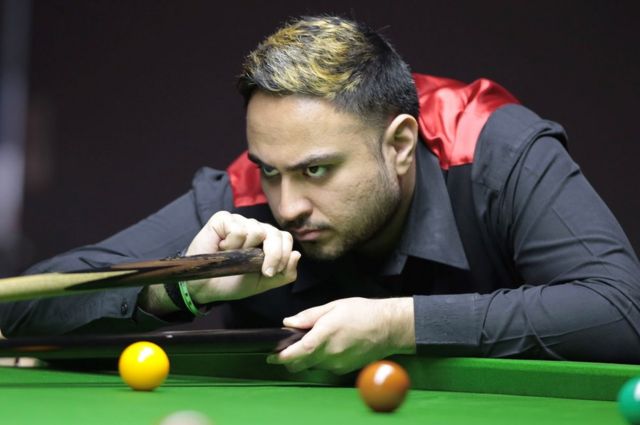 An Asian snooker player wearing a smart black shirt and waistcoat with a velvety red back leans over the green baize of a snooker table. He's concentrating hard as he holds a cue in one hand and a rest with his other. A yellow, brown and green ball can be seen on the table.