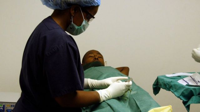 Paciente acostado en una cama mientras una trabajadora de la salud prepara el área para la circuncisión.