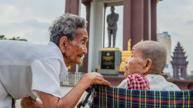 Bun Sen agacha-se perto de Bun Chea, que está em uma cadeira de rodas; ambas aparecem em frente a estátua de figura histórica na capital cambojana, Phnom Penh