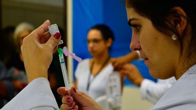 Profissional preparando seringa para vacinação com mulher sendo vacinada ao fundo