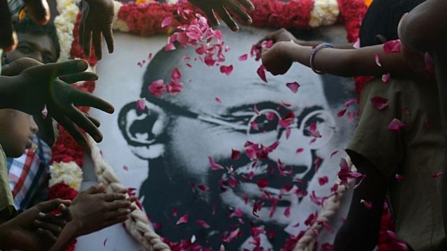 Mãos jogando pétalas de flores em retrato de Mahatma Gandhi