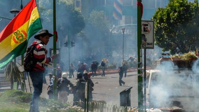 Homem com bandeira da Bolívia em meio a protesto, cercado de gás lacrimogêneo