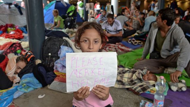 Rebecca de Siria, una niña migrante sostiene un trozo de papel 'Please Merkel Help Me' (Por favor, ayúdame Merkel) mientras su familia espera el transporte a Alemania, en la estación de tren de Budapest Keleti, en 2015.
