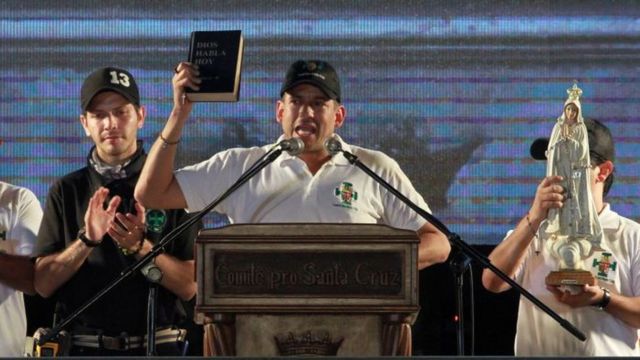 Luis Fernando Camacho en un mitín con la biblia en la mano mientras alguien sostiene a su lado una virgen