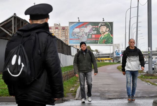 Двое мужчин идут по улице на фоне плаката, призывающего добровольно идти на войну