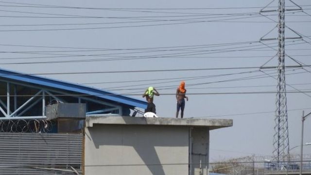 Dois presos com camisas na cabeça aparecem em pé em cima de telhado de penitenciária