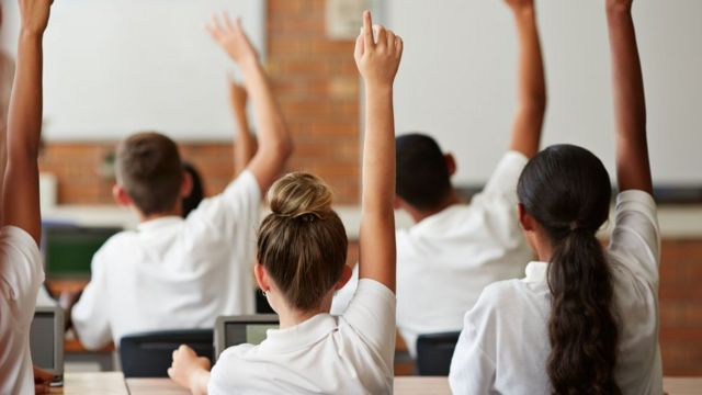 Estudantes de costas, sentados, com os braços levantados em sala de aula