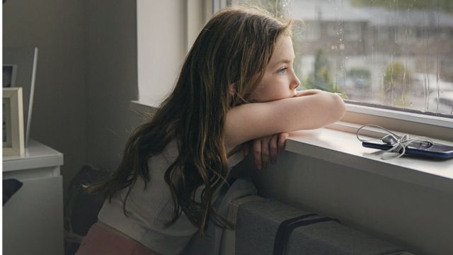 Une fille blanche aux cheveux longs, assise sur les toilettes, regarde par la fenêtre d'un air triste.