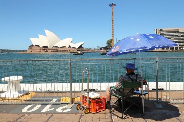 คนนั่งริมน้ำกางร่มลายธงชาติออสเตรเลีย เห็นวิวโรงละครโอเปรา