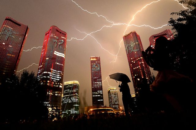 A bolt of lightning crosses the sky as people look at buildings displaying a light show on the eve of the 100th anniversary of the Chinese Communist Party in Beijing