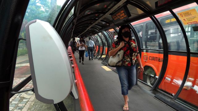 Pedestres com máscara caminham em estação-tubo, com ônibus atrás, à luz do dia