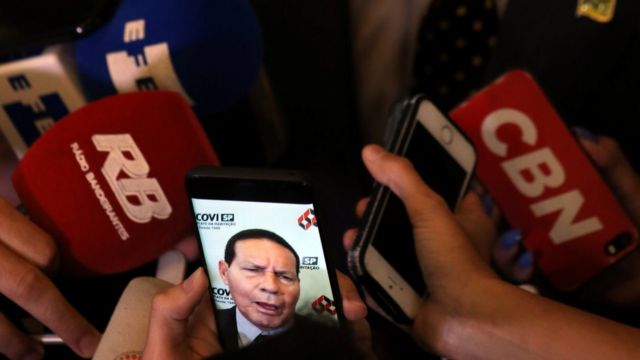 Imagem de Mourão aparece em celular em meio a microfones de diversos veículos de imprensa