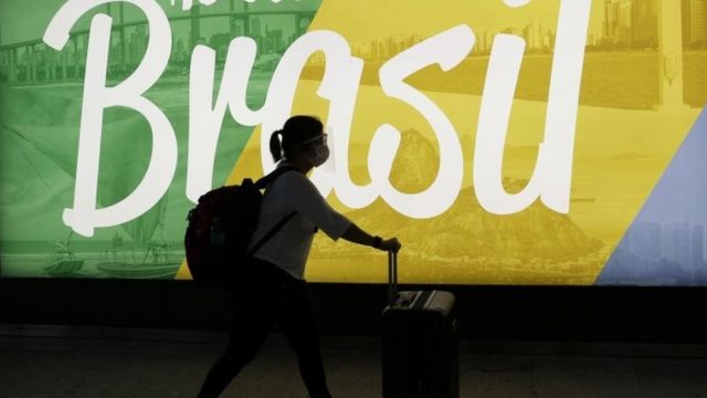 Mulher caminha com mala em frente a propaganda do Brasil