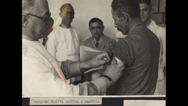 Foto em preto e branco mostra homem, de costas, sendo vacinado por outro homem em sala