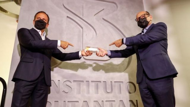 O governador de São Paulo, João Doria, e o diretor do Butantan, Dimas Covas, aparecem de máscara em frente a painel com inscrição 'Instituto Butantan' segurando juntos embalagem da Coronavac