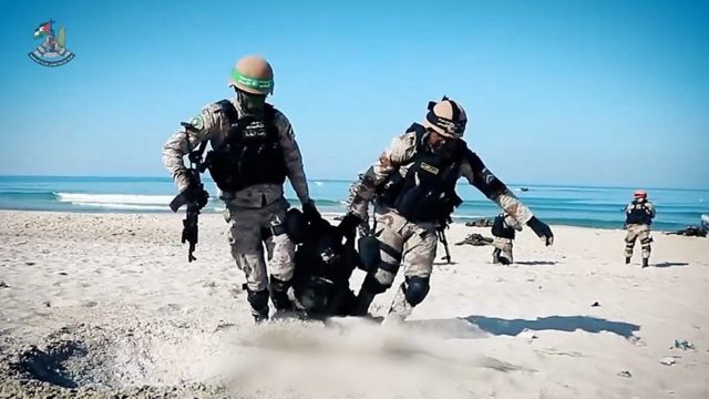 训练中，两名身着迷彩服的男子在沙滩上拖着另一名男子