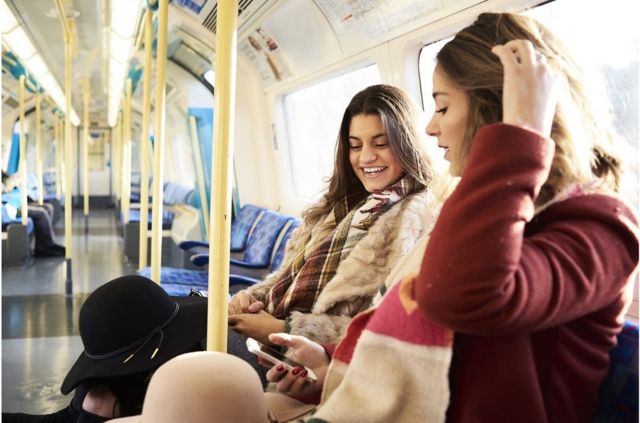 Two women look at mobile phones and talk (profile photo) in the London Underground.