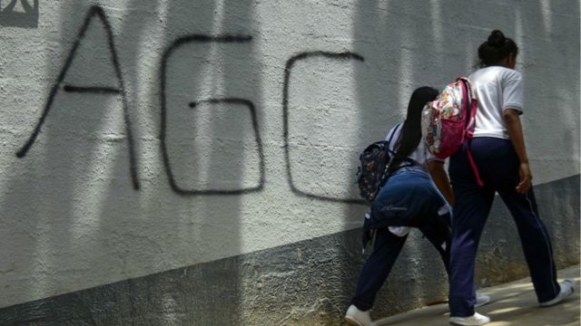 Duas jovens de costas e com mochilas passam por pichação no muro, escrito "AGC"