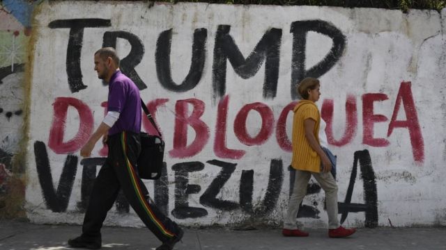 Un hombre y una mujer pasan frente a un muro con una pancarta que lee: "Trump desbloquea Venezuela"